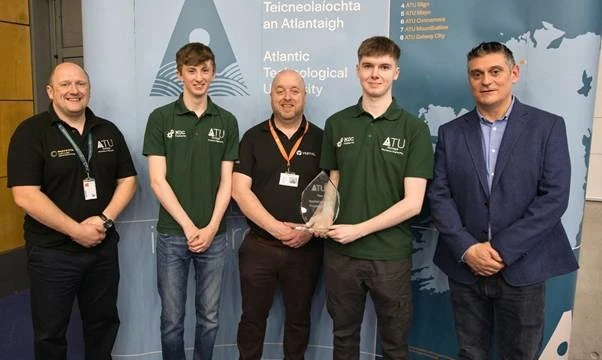 Photograph of five people standing indoors in front of a backdrop featuring Atlantic Technological University branding and a map. One person holds a glass award.