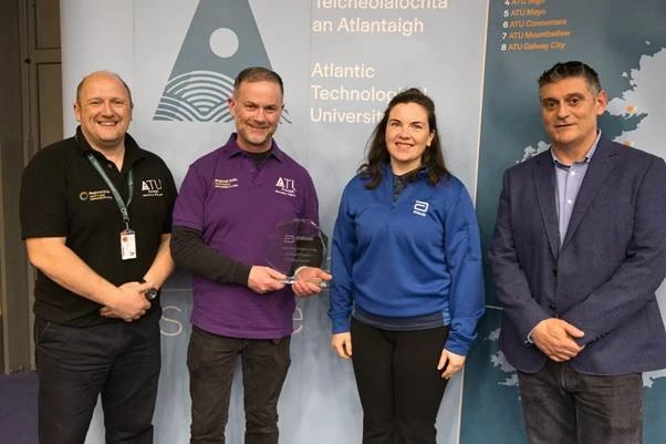 Photograph of four people standing indoors in front of a backdrop featuring Atlantic Technological University branding and a map. One person holds a glass award.