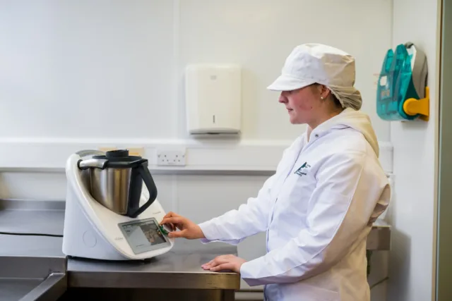 Photograph of a person in a white lab coat and hairnet operating a kitchen appliance with a digital touchscreen on a stainless steel counter.