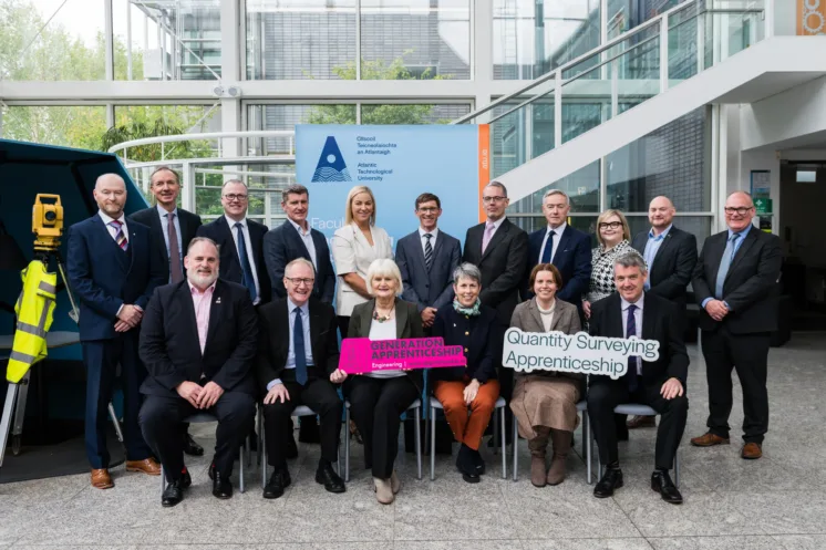 Photograph of a group of professionals posing indoors at the launch event, with two seated individuals holding signs reading "Generation Apprenticeship" and "Quantity Surveying Apprenticeship." Background includes a large banner with the ATU logo and text, and the group is dressed in business attire.