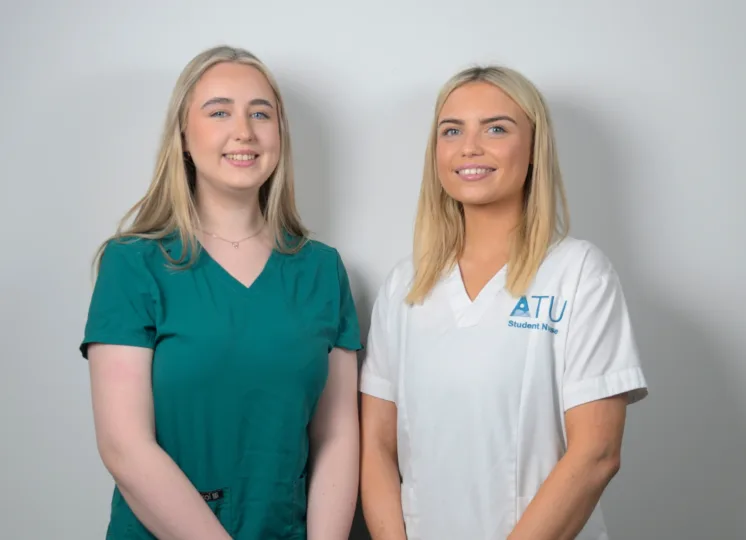 Photograph of two female students standing side by side against a plain background, wearing medical scrubs. One student wears green scrubs, and the other wears white scrubs with "ATU Student Nurse" printed on the chest.