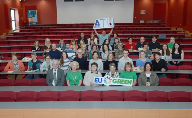 Photograph of a group of people seated in a lecture hall with tiered red benches, holding signs reading "ATU" and "EU GREEN."