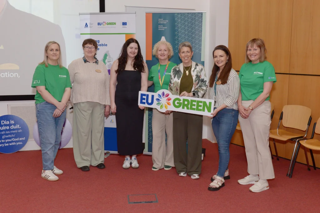 Photograph of seven people standing indoors in front of promotional banners, with three individuals wearing green shirts and holding a sign that reads "EU GREEN."