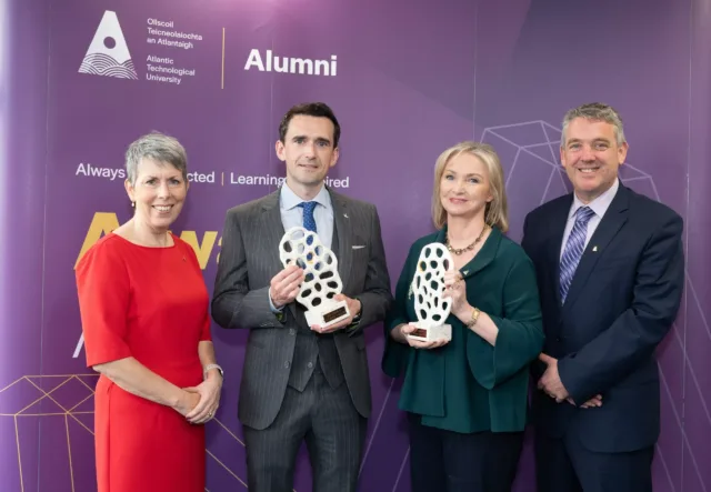 Four people stand in front of a purple backdrop holding award. Two individuals in the centre each hold a white, patterned trophy on a base while the others stand alongside.