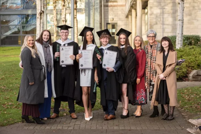 Group of nine people standing outdoors on a paved path in front of a modern building and trees, with four individuals wearing graduation gowns and mortarboards and holding certificates. The group is posed together for a photograph in daylight.