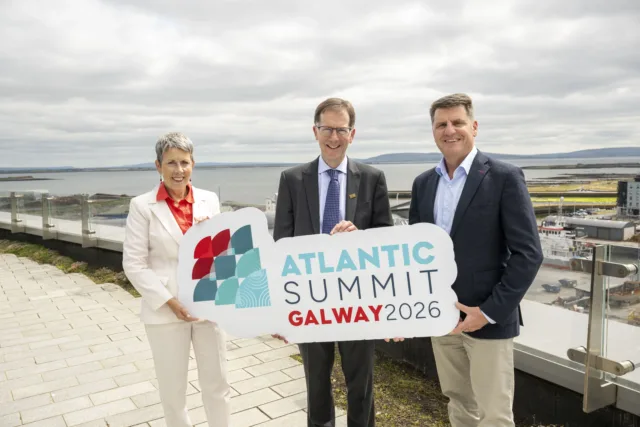 Photograph showing three people standing outdoors on a terrace overlooking a coastal area, holding a sign that reads "Atlantic Summit Galway 2026." The individuals are dressed in business attire.