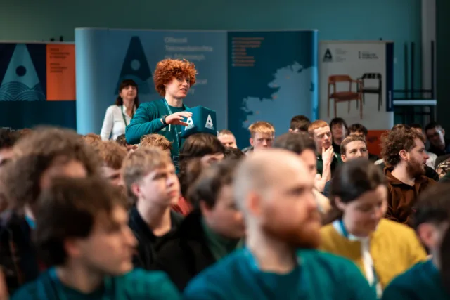 Photograph of a conference with a diverse audience seated and focused on a speaker or event. A person with curly red hair stands holding a microphone cube with an ATU logo.