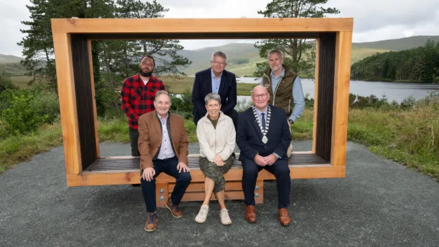 Photograph of six people posing outdoors on and around a large wooden frame bench with a scenic lake and hills in background. Group includes three seated individuals in front and three standing behind.