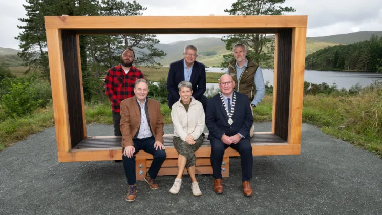 Photograph of six people posing outdoors on and around a large wooden frame bench with a scenic lake and hills in background. Group includes three seated individuals in front and three standing behind.
