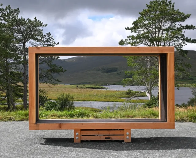 Photograph of a large wooden frame sculpture positioned outdoors, perfectly framing a scenic landscape of a river, grass fields, trees, and distant hills under a cloudy sky. The frame serves as a natural picture window.