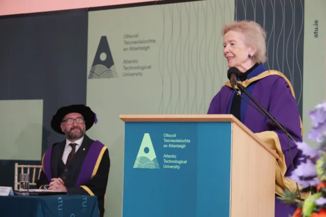 Photograph of conferring ceremony, at Atlantic Technological University, showing a woman in academic regalia speaking at a podium with the university logo and name displayed behind her. A man in similar academic attire is seated at a table nearby, attentively listening.