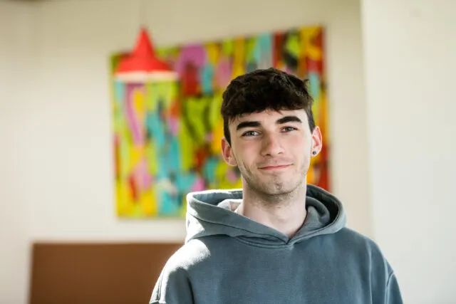 Male student in grey hoodie, standing in front of colourful picture