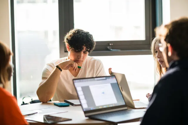 Male student in focus, working on a group project with other students with open laptop on the desk