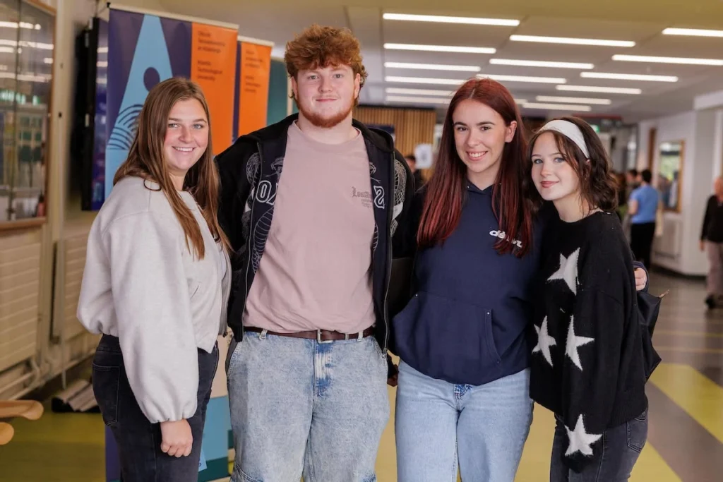 Photograph showing four smiling young people posing together in a brightly lit university corridor.