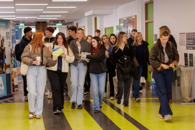 Photograph shows a large group of students walking through a brightly lit college corridor. Students carry books, drinks and bags, while green-and-black striped flooring, green-framed doors and directional signs create a busy campus scene.