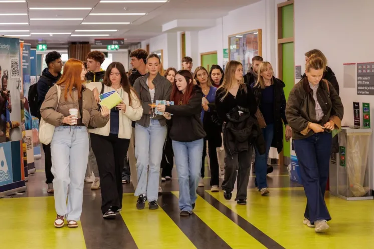 Photograph shows a large group of students walking through a brightly lit college corridor. Students carry books, drinks and bags, while green-and-black striped flooring, green-framed doors and directional signs create a busy campus scene.
