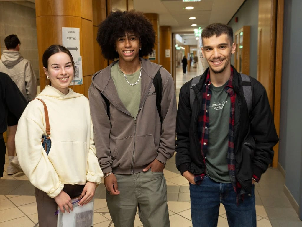 Photograph showing three smiling young adults posing in a university corridor. A small wall sign, tiled floor, backpacks and casual clothing provide campus context,