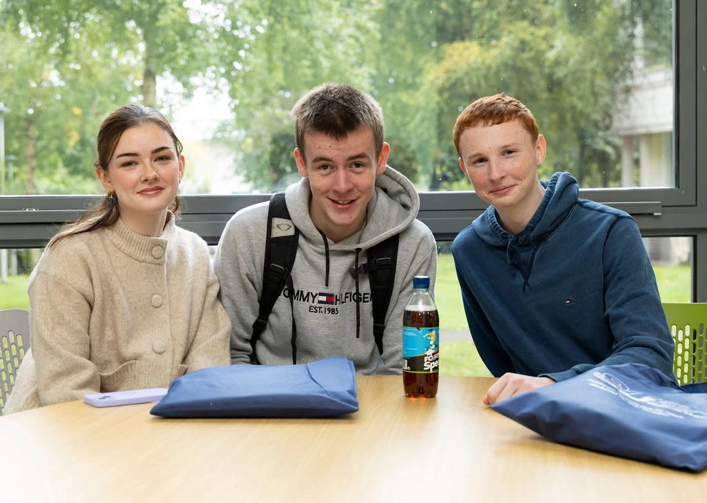 Photograph shows three young people seated around a table in a bright room at student induction. Blue tote bags, smartphone, a bottled drink sit on the table. A large windows with greenery is the background.
