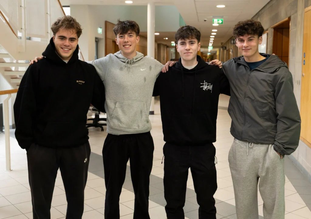 Photograph of four young men standing together in a bright university corridor, smiling with arms around one another. They wear casual black, grey and olive clothing, with tiled flooring, stairway and hallway visible behind them.