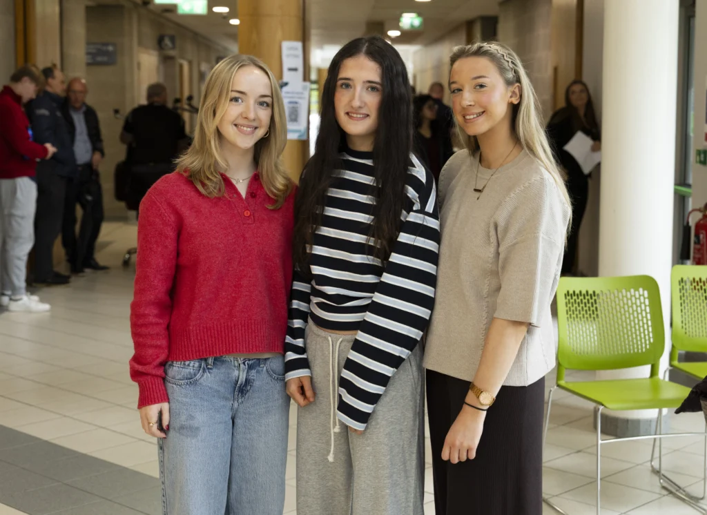 Photograph of three young women smiling together in a public-building corridor. Red jumper, striped top and beige knitwear contrast with green chairs, tiled flooring and people in background