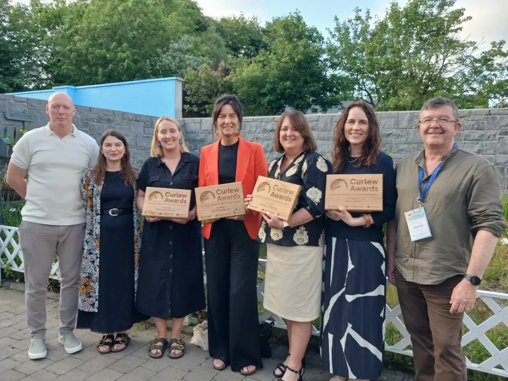 Six people standing outdoors in front of a stone wall and garden area, with four people holding wooden plaques that read “Curlew Awards”. Trees and a light-blue structure are visible in the background.