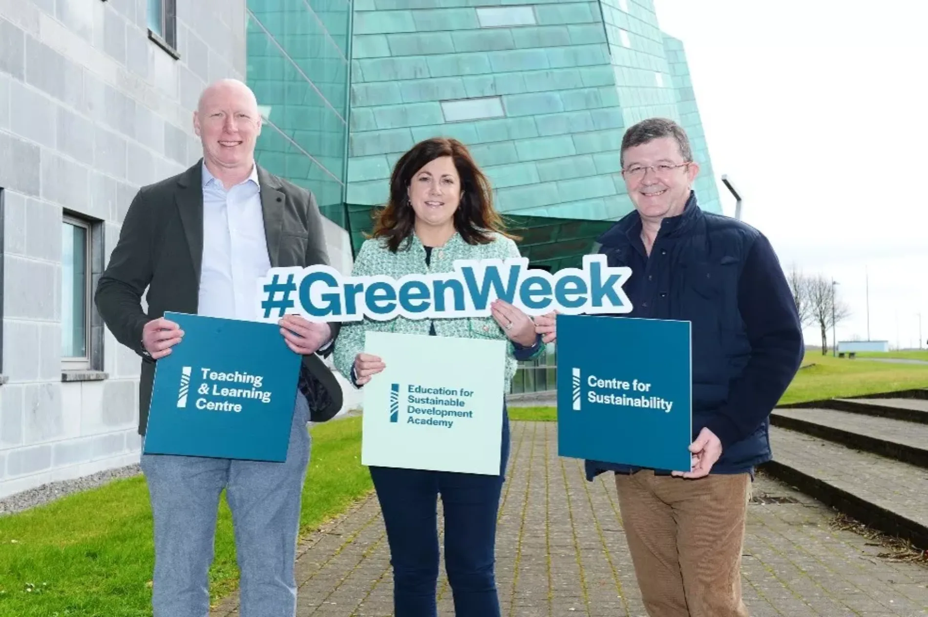 Three people standing outdoors in front of a modern building, holding signs and folders. The signs display “#GreenWeek”, “Teaching & Learning Centre”, “Education for Sustainable Development Academy”, and “Centre for Sustainability”.